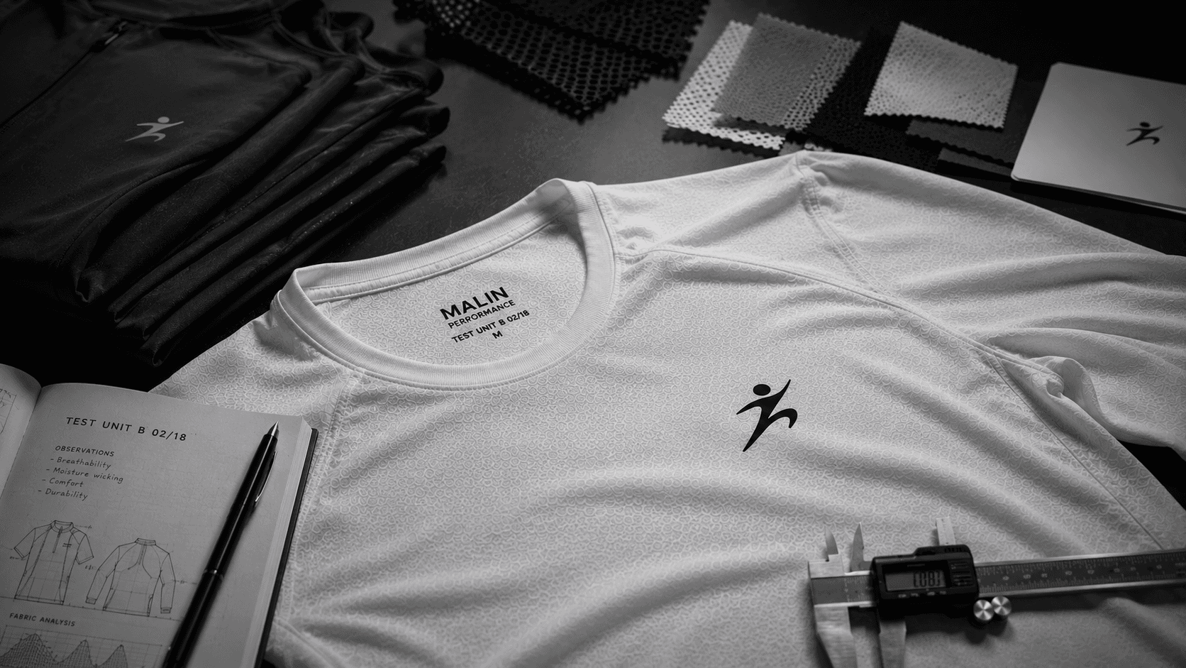 A white Malin performance tee laid out with fabric swatches, a spec notebook and calipers